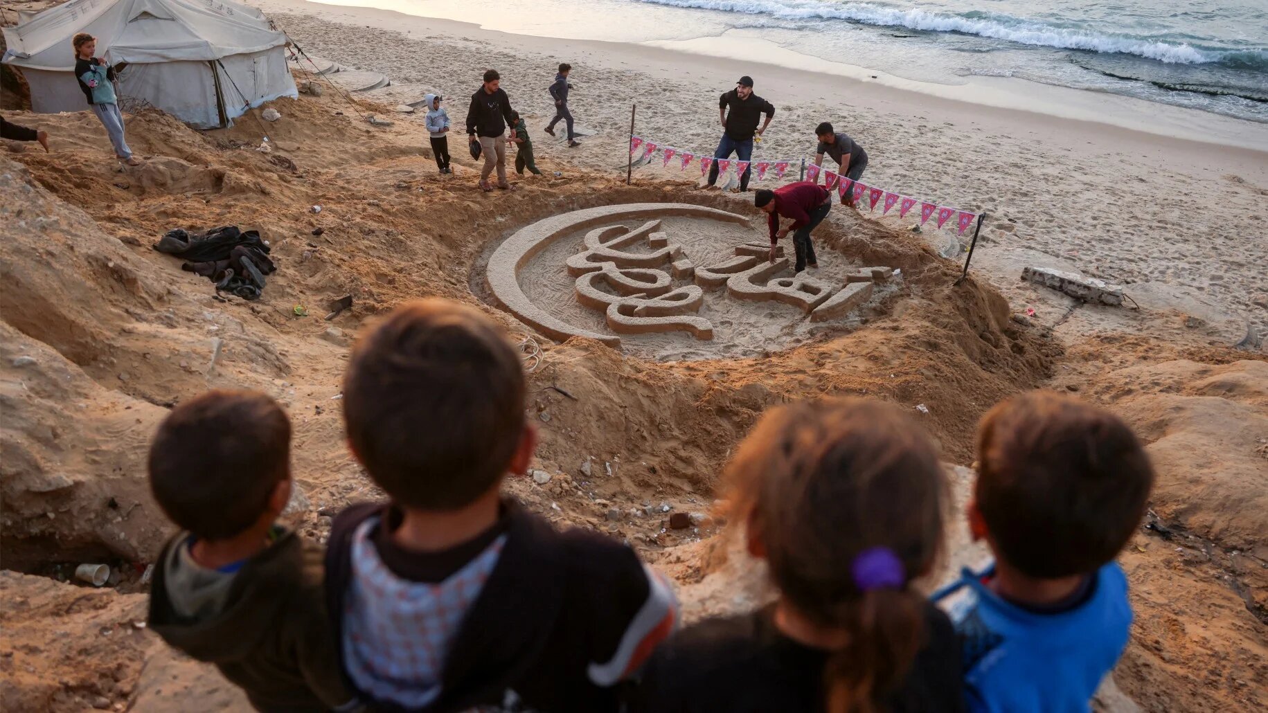 Children stand on a dune above a sand sculpture with the greeting message "Welcome, Ramadan" created by Palestinian artist Yazeed Abu Jarad, along a beach in Khan Yunis on 17 February 2026, a day ahead of the start of Ramadan (Bashar Taleb/AFP)